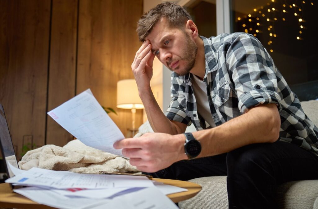 Worried man reviewing bills at home while holding his head and looking at paperwork.