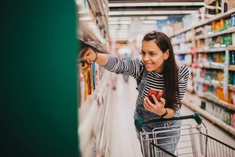Smiling woman with a shopping trolley reaching for a product in a supermarket aisle