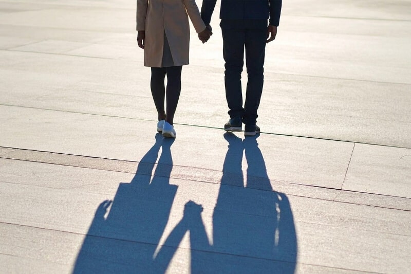 Couple walking hand in hand across a sunlit plaza, casting long shadows ahead