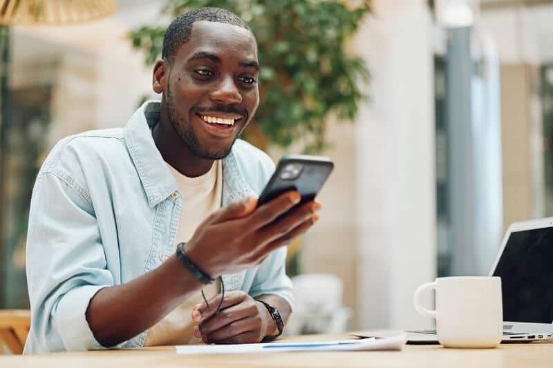 Smiling man looking at his phone at a desk with a laptop and coffee mug nearby