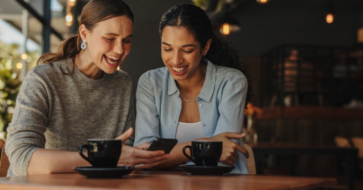Two women laughing together over a phone at a café table with coffee cups